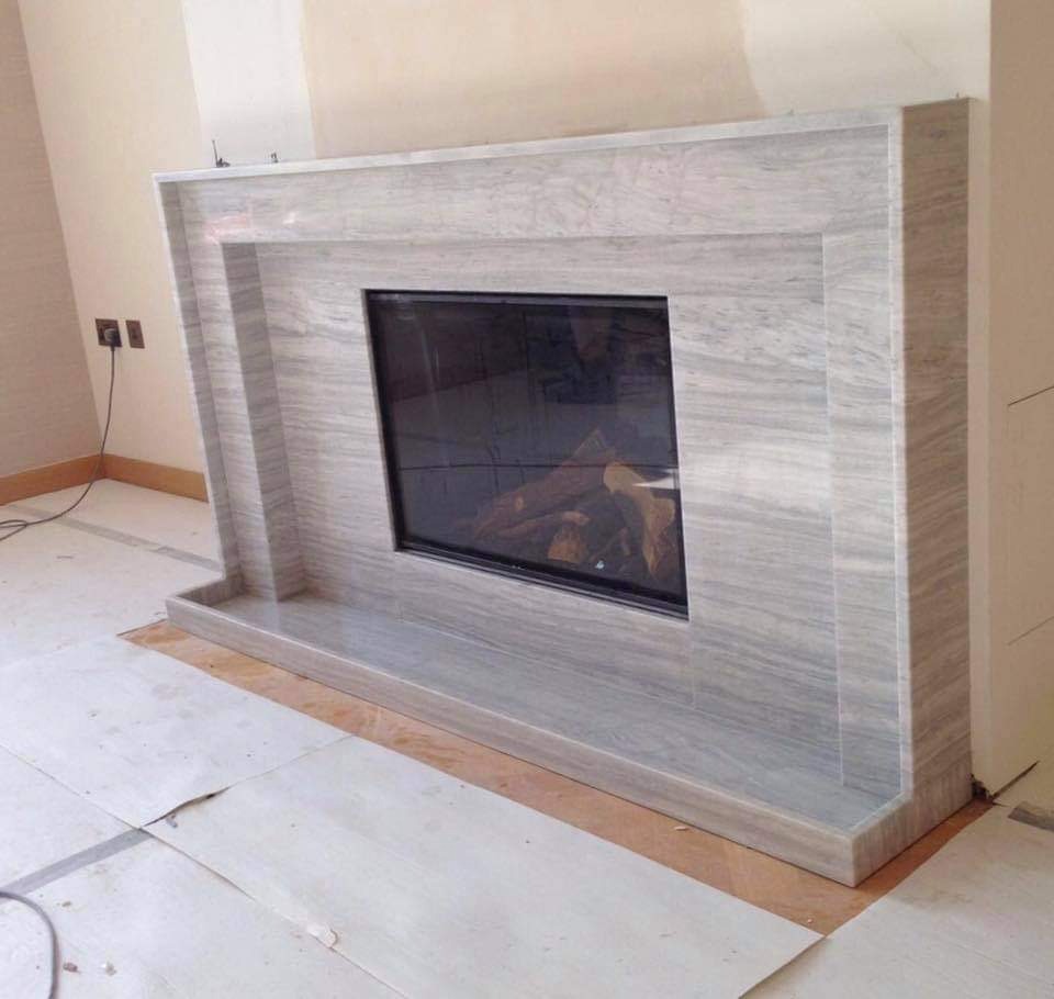 Wykończenie kominka z szarego kamienia z widocznym wkładem kominkowym, częściowo zabezpieczone podłoże w trakcie prac montażowych.