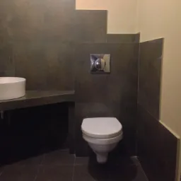 Wykończona łazienka z ciemnymi płytkami i białym WC oraz umywalką. Nietypowe ułożenie płytek na ścianie. Nowoczesny design i minimalizm.