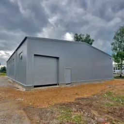 Szara hala przemysłowa z bramą rolowaną i małymi oknami, widok z zewnątrz w pochmurny dzień, teren wokół wysypany żwirem.