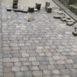 Układanie szarej kostki brukowej na podjeździe, widoczne stosy kostki przygotowane do ułożenia, oraz fragment ułożonej nawierzchni z kratką odpływową.