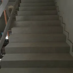 Schody wyłożone szarymi płytkami ceramicznymi, widok z dołu, z widocznym fragmentem drewnianego rusztowania po lewej stronie i niedokończonym wykończeniem ściany po prawej.