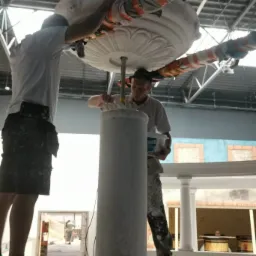 Dwóch mężczyzn montuje i wykańcza ozdobną kolumnę z elementami sztukaterii w przestronnym wnętrzu. Jeden z nich trzyma pojemnik z farbą.