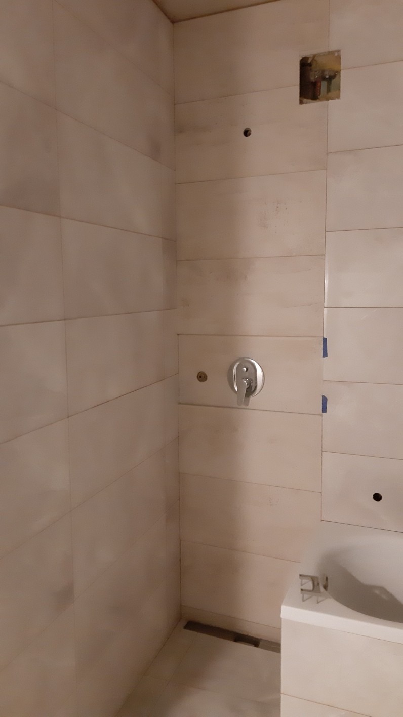 Fragment łazienki w trakcie remontu z beżowymi, poziomymi płytkami, widoczne otwory na armaturę, bateria prysznicowa i niezabezpieczony otwór w ścianie.