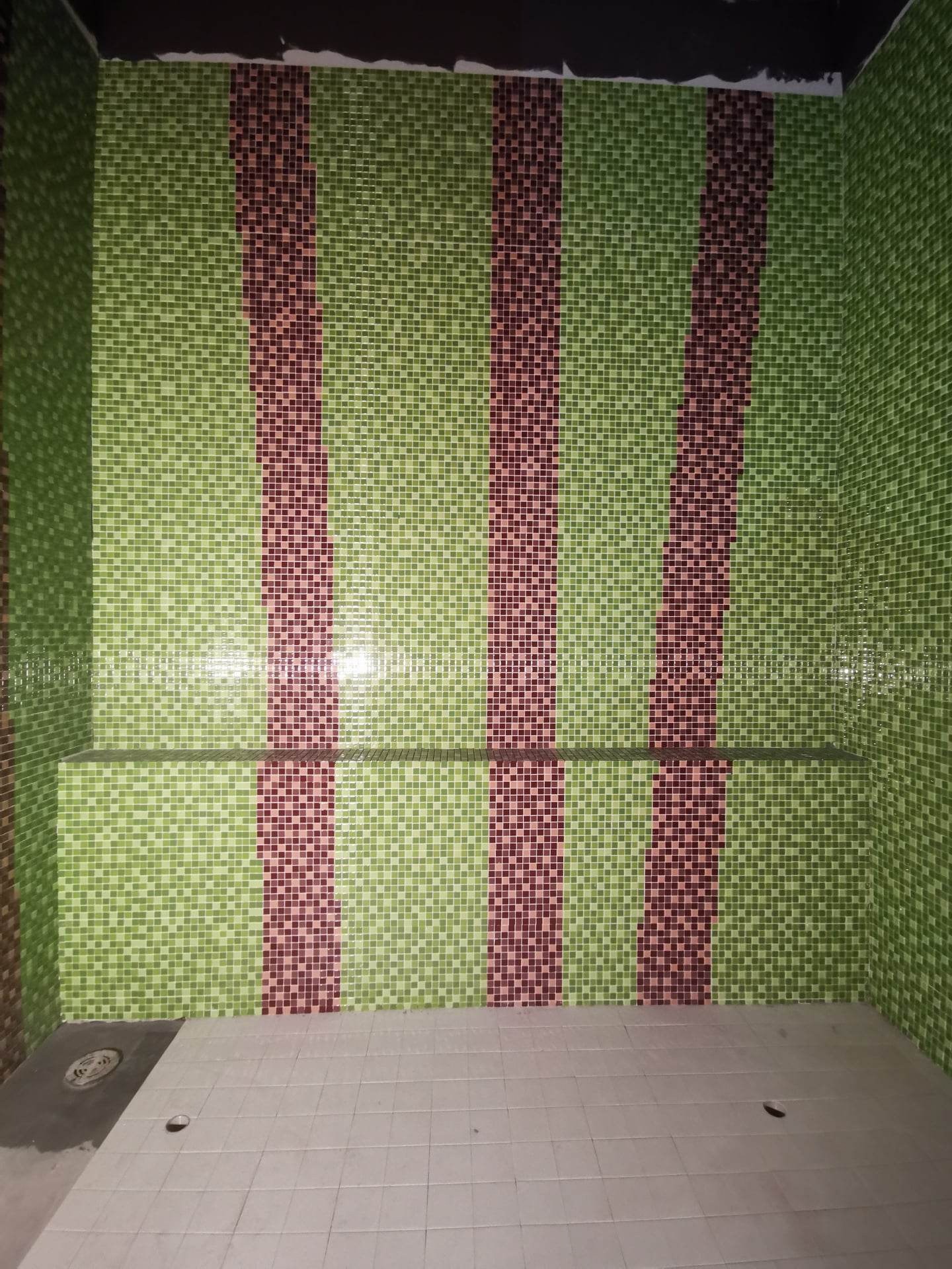 Ściana łazienki wyłożona mozaiką w odcieniach zieleni i bordo, z półką na środku i odpływem liniowym w podłodze, widoczne niedociągnięcia na łączeniu ściany z sufitem.