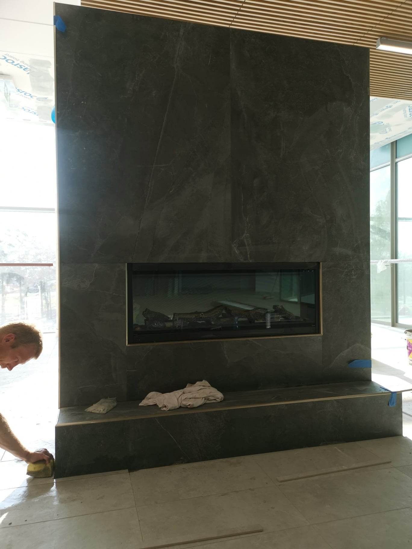 Obróbka kamieniarska wokół kominka z ciemnych płyt imitujących marmur, widoczny fragment twarzy i dłoni pracownika czyszczącego powierzchnię wilgotną gąbką.