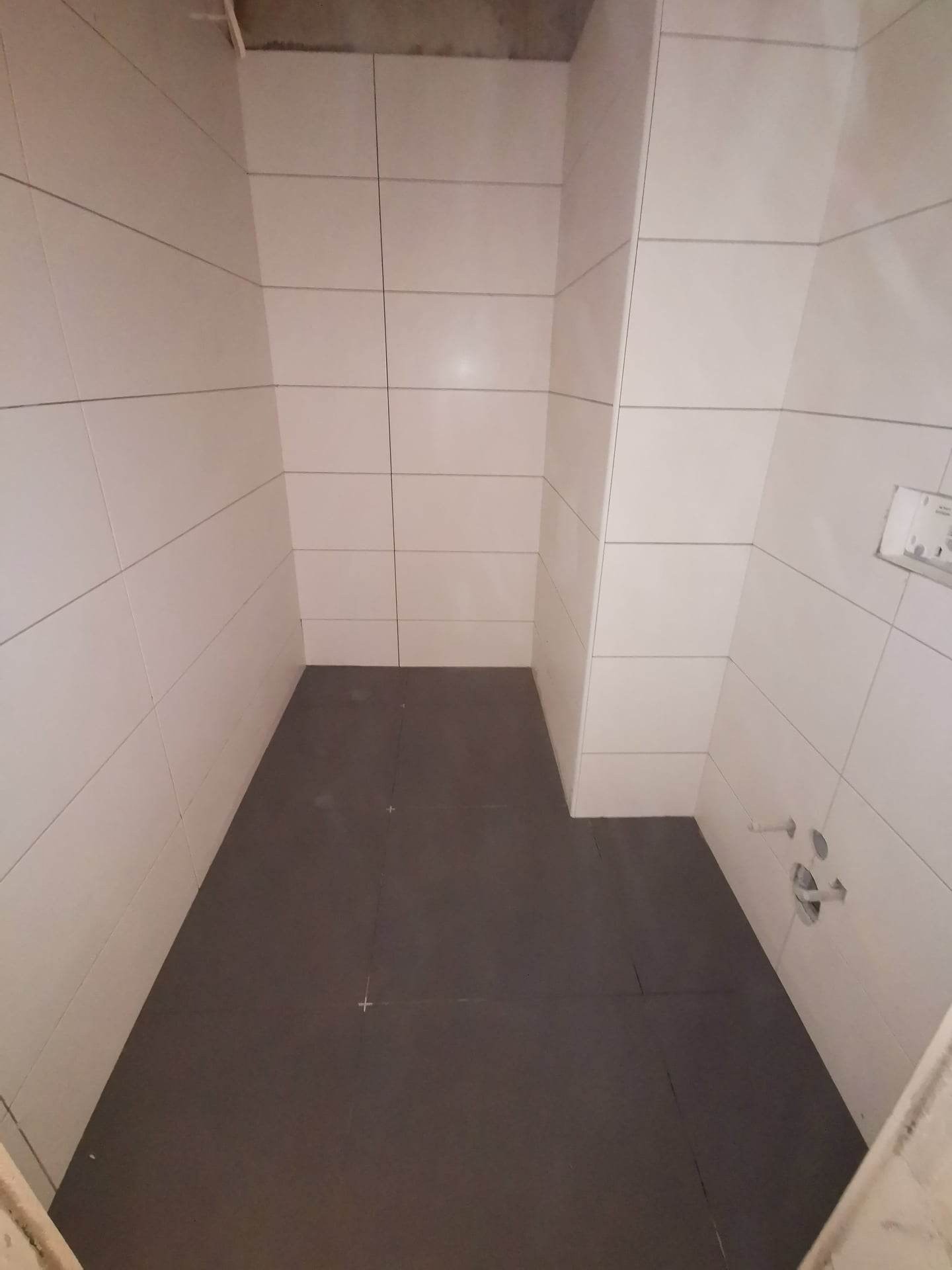 Wnętrze łazienki w trakcie prac wykończeniowych, widoczne białe płytki na ścianach i ciemna, zabezpieczona folią podłoga z zaznaczonymi krzyżykami, surowy betonowy sufit.