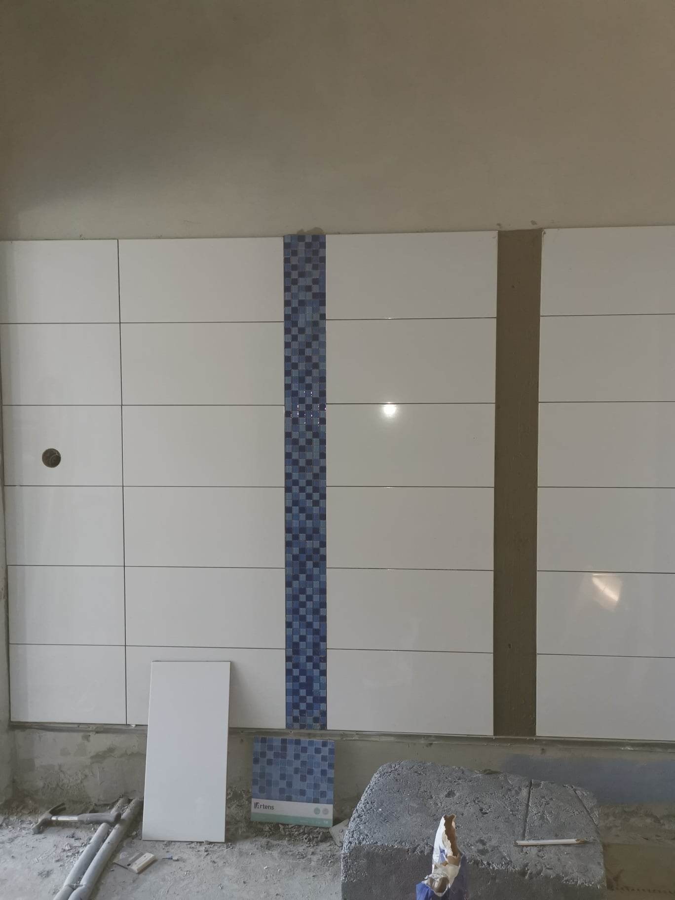 Ściana w trakcie układania białych płytek z dekoracyjnym pasem niebieskiej mozaiki, widoczne przygotowane płytki i narzędzia glazurnicze.