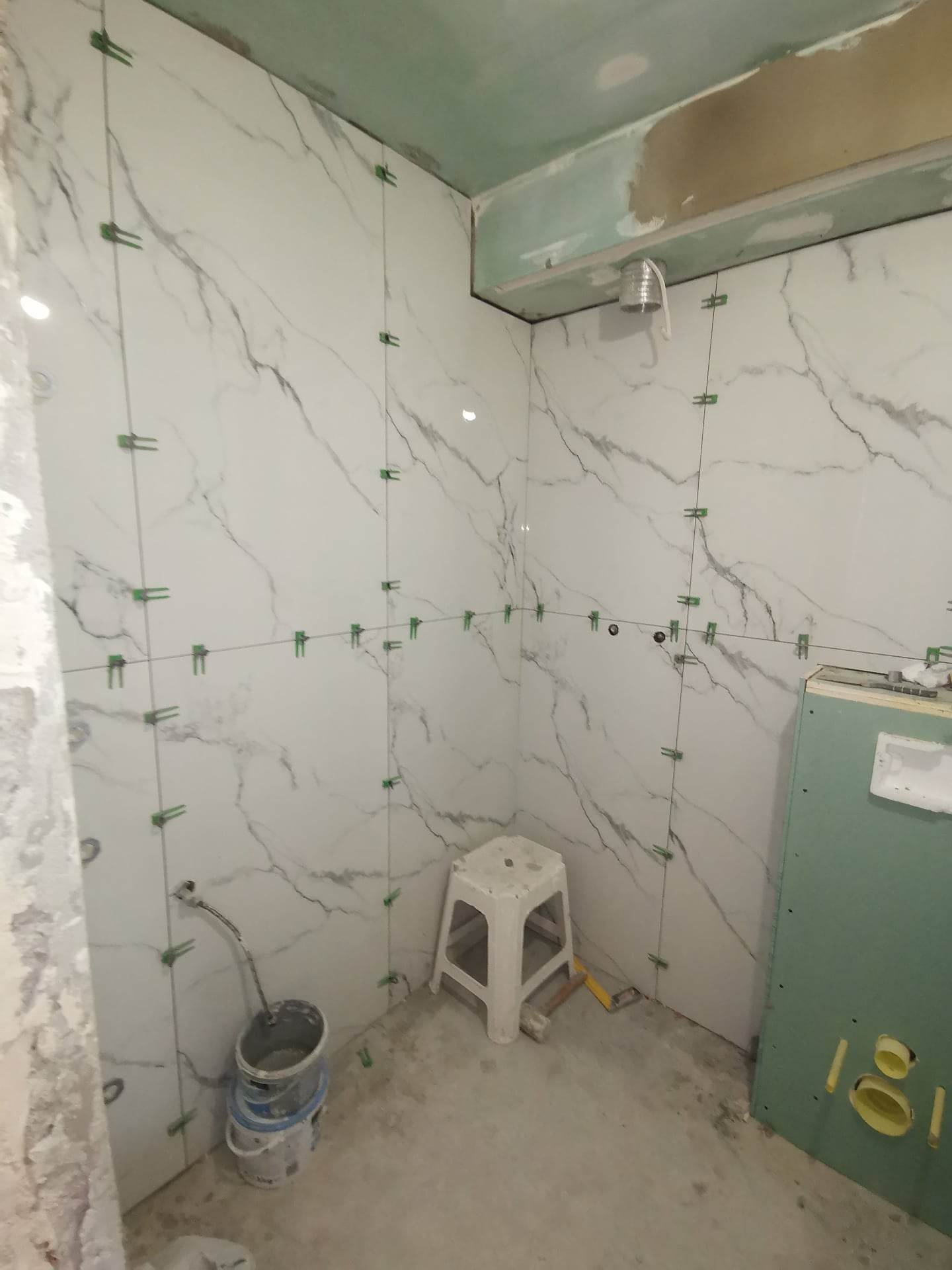 Białe płytki imitujące marmur, z zielonymi krzyżykami dystansowymi, w trakcie układania na ścianie łazienki; widoczne wiadro z zaprawą, plastikowy taboret i fragment zabudowy z płyty...