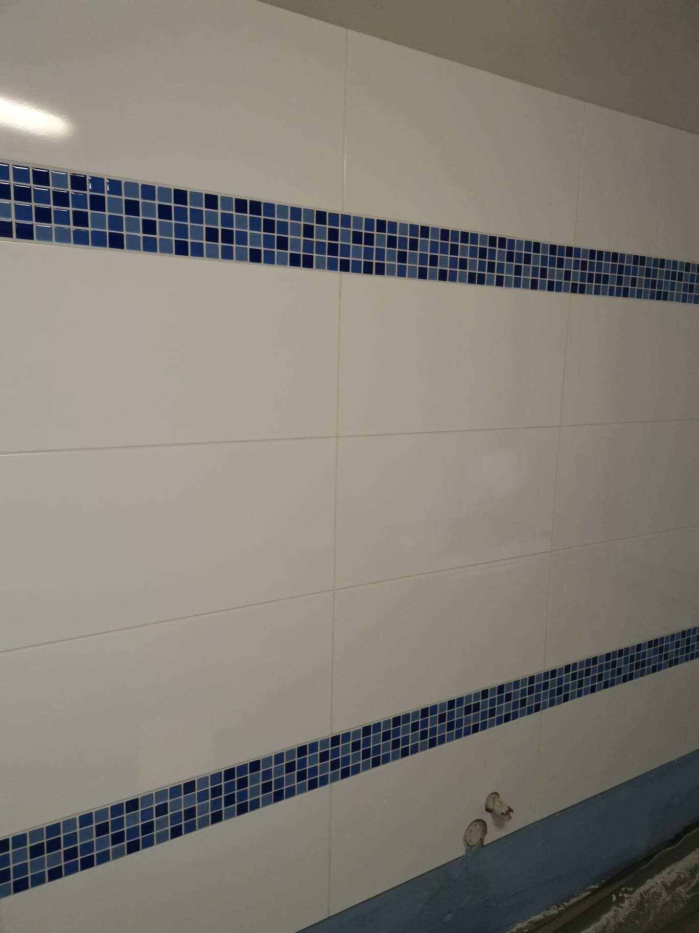 Ściana wyłożona białymi, prostokątnymi płytkami z dwoma poziomymi pasami dekoracyjnej, niebieskiej mozaiki, widoczne wystające rury instalacji hydraulicznej przy podłodze.