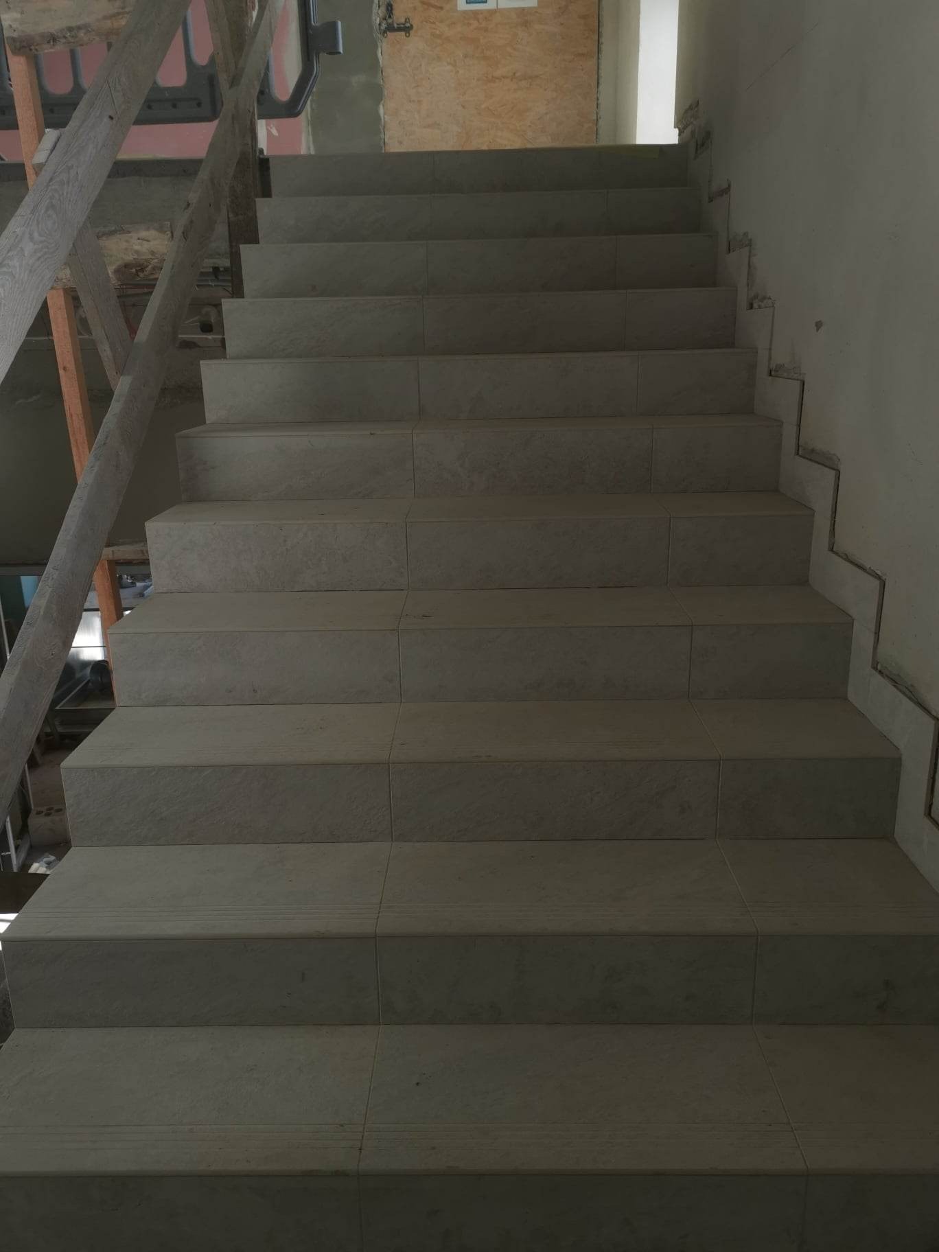 Schody wyłożone szarymi płytkami ceramicznymi, widok z dołu, z widocznym fragmentem drewnianego rusztowania po lewej stronie i niedokończonym wykończeniem ściany po prawej.