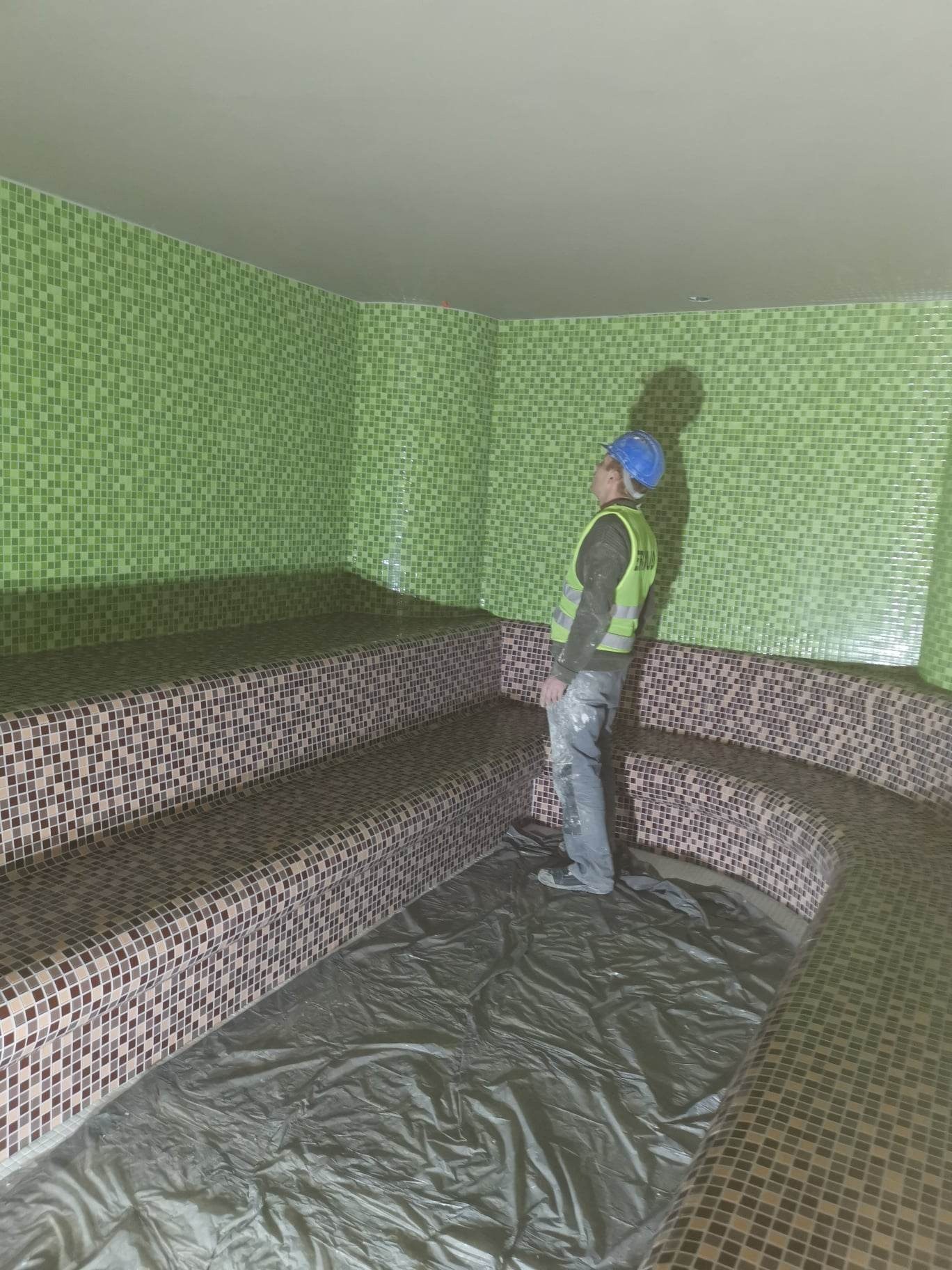 Pracownik w kasku i kamizelce odblaskowej ogląda zieloną ścianę i mozaikowe ławy w saunie, podłoga zabezpieczona folią.