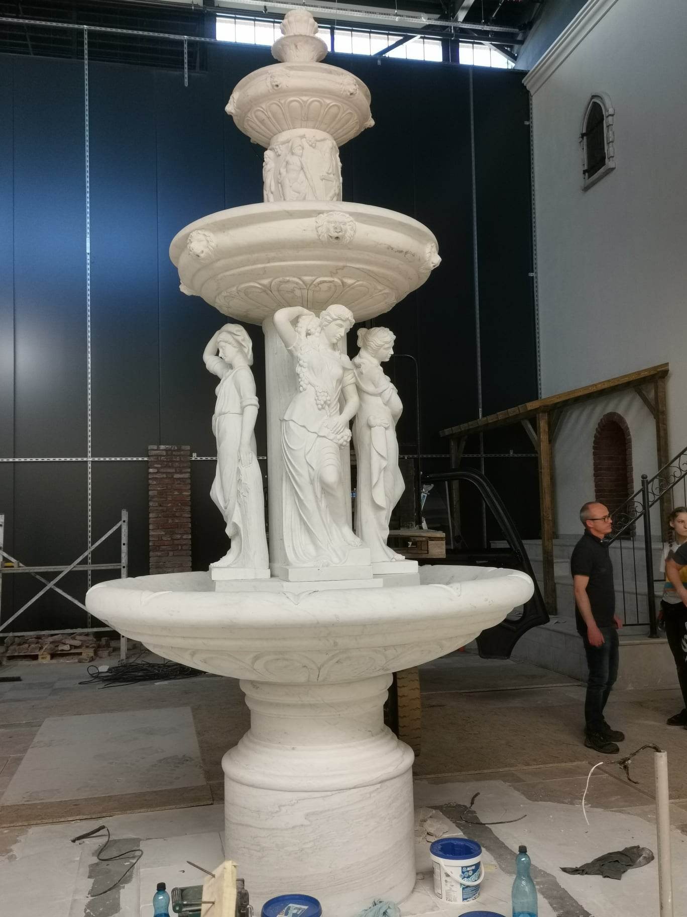 Wielopoziomowa, biała fontanna z rzeźbami kobiet w klasycznym stylu, ustawiona w surowym wnętrzu z widocznymi rusztowaniami i elementami budowlanymi, sugerująca etap montażu lub wykończenia.