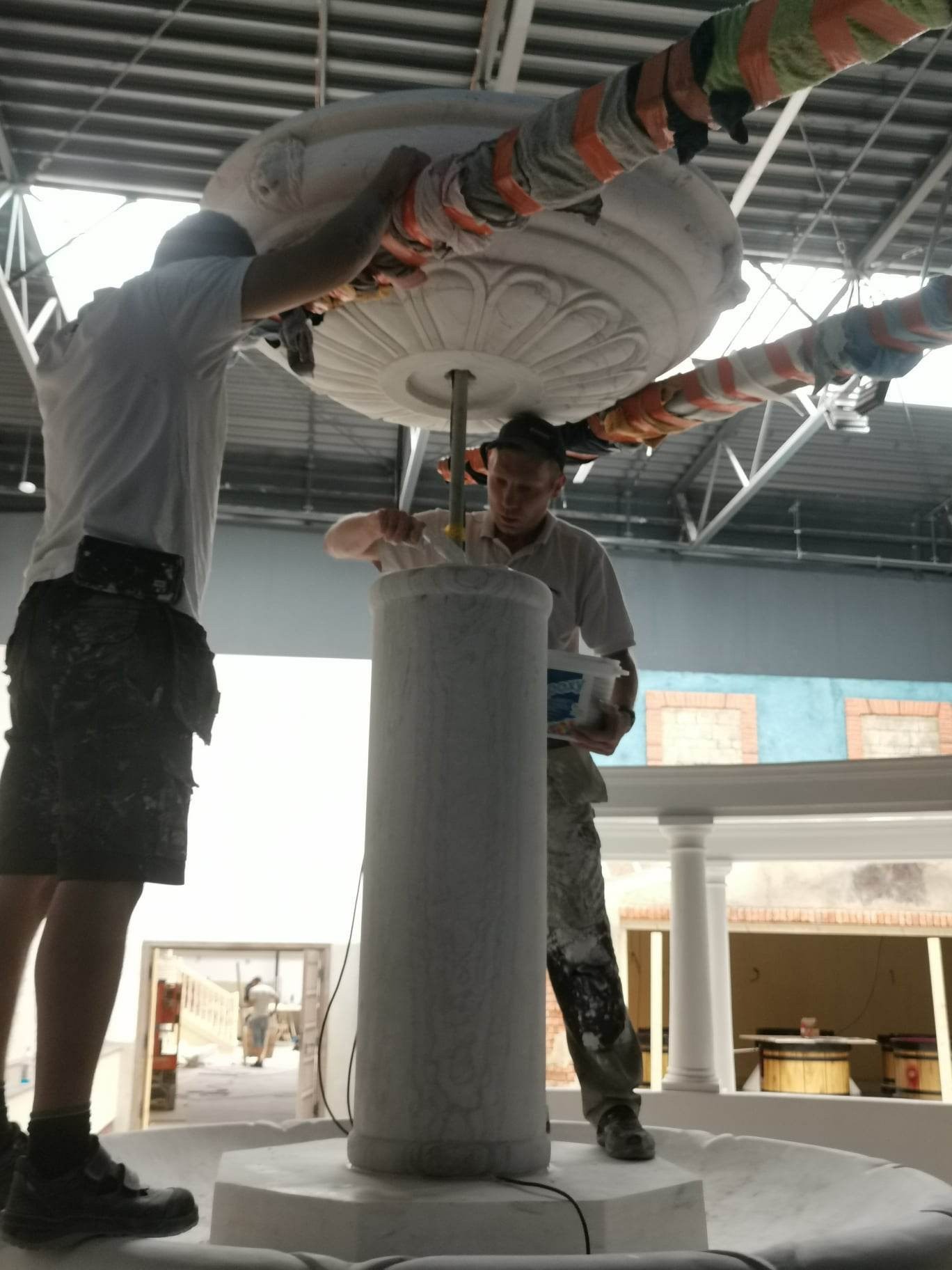 Dwóch mężczyzn montuje i wykańcza ozdobną kolumnę z elementami sztukaterii w przestronnym wnętrzu. Jeden z nich trzyma pojemnik z farbą.