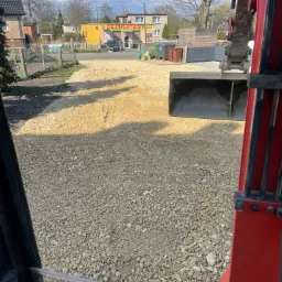 Widok z kabiny koparki na teren pokryty drobnym kruszywem; w tle budynki i drzewa w słoneczny dzień.