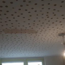 Przygotowanie sufitu do montażu płyt karton gips: nawiercony sufit z widocznymi otworami, drabina oparta o okno zabezpieczone folią malarską, lampa wisząca w rogu.