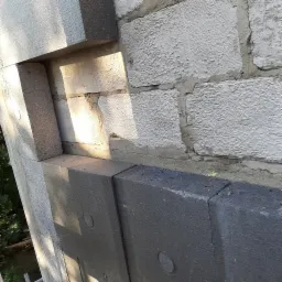 Fragment ściany budynku z widoczną warstwą ocieplenia z grafitowego styropianu, częściowo zakrywającego białe bloczki betonowe, z widocznymi śladami zaprawy.