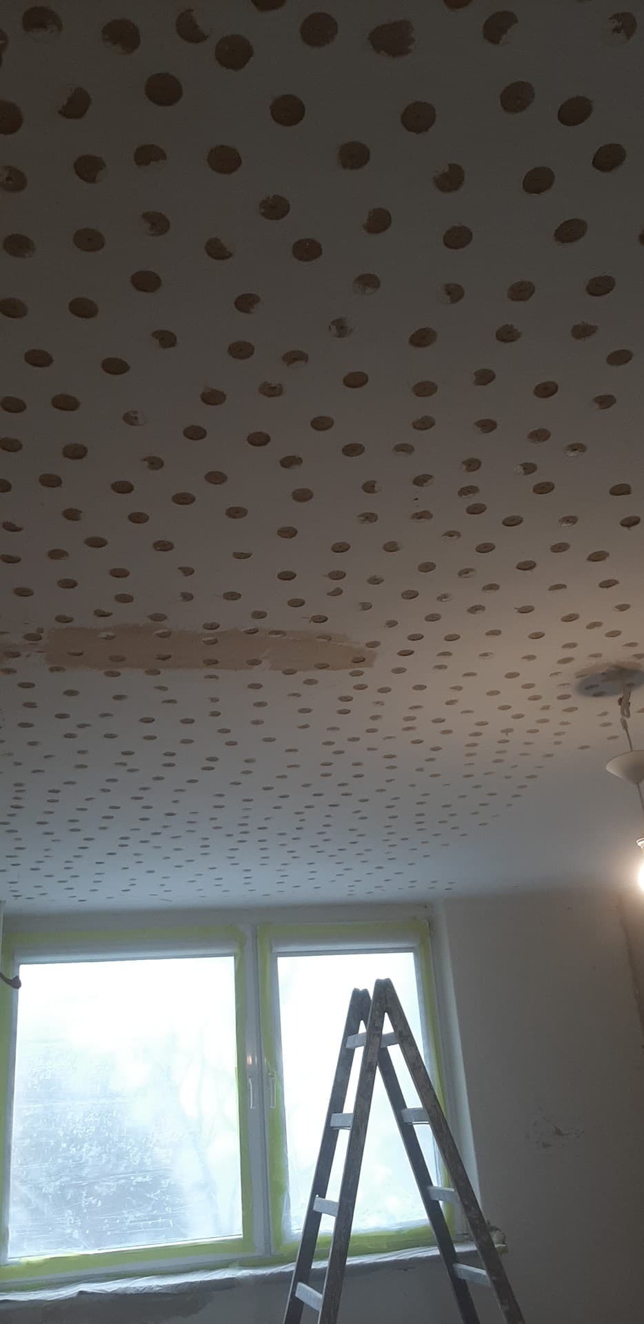 Przygotowanie sufitu do montażu płyt karton gips: nawiercony sufit z widocznymi otworami, drabina oparta o okno zabezpieczone folią malarską, lampa wisząca w rogu.