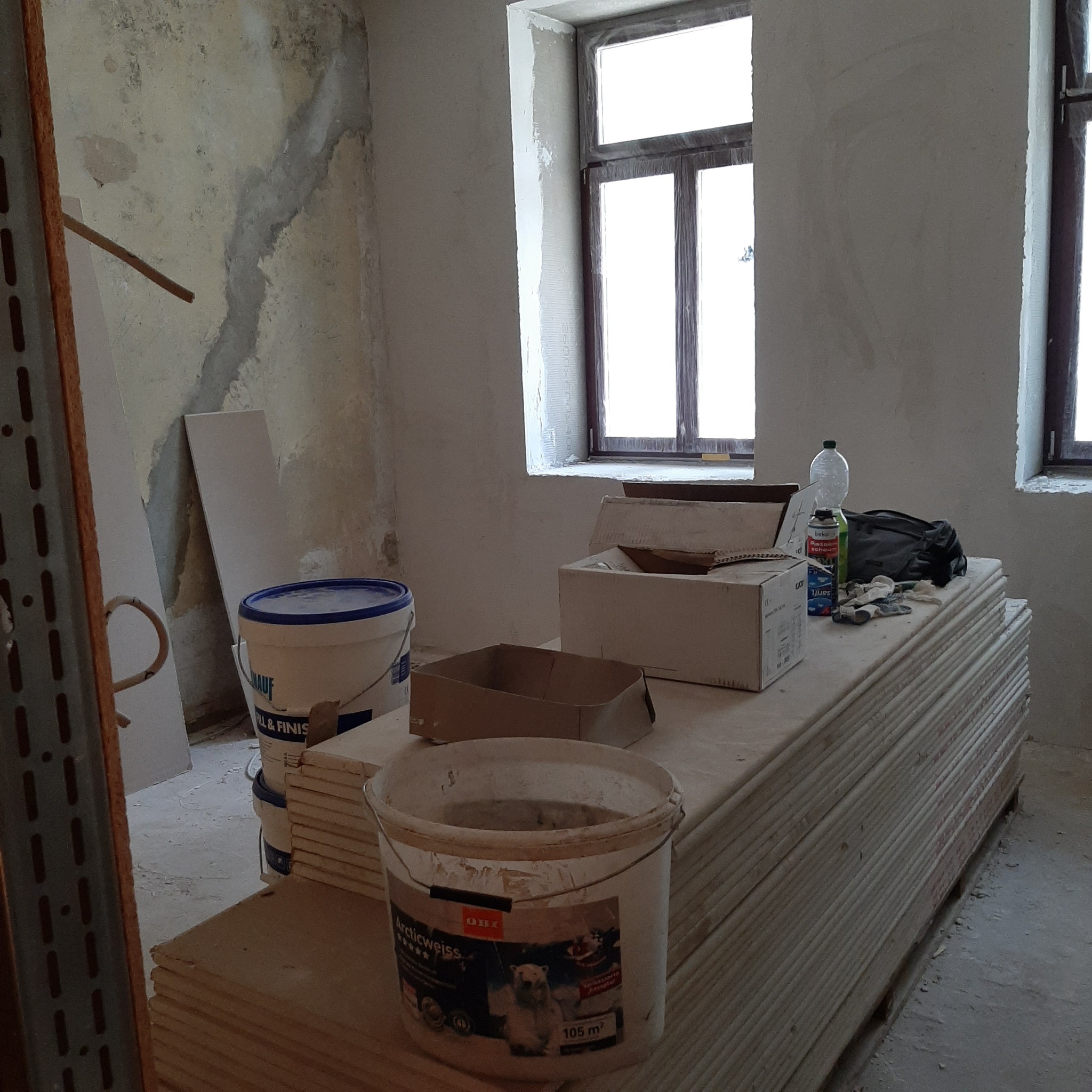 Stos płyt kartonowo-gipsowych w surowym wnętrzu, obok wiadra z farbą, kartony i narzędzia, ściany w trakcie przygotowania do malowania, widoczne ślady po starych warstwach wykończeniowych.
