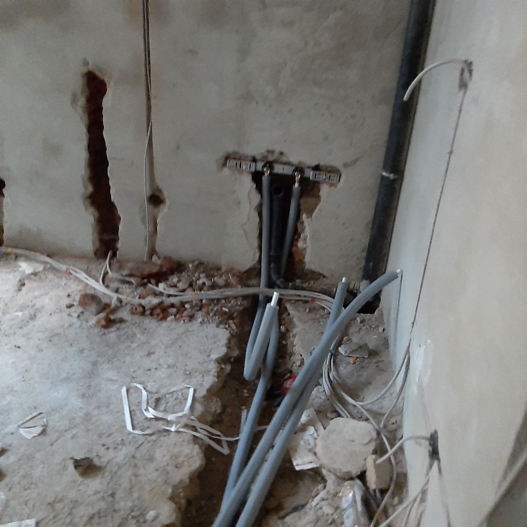 Surowy stan ściany z widocznymi przewodami instalacji wodnej i elektrycznej, fragmenty gruzu na podłodze, przygotowanie do prac wykończeniowych.