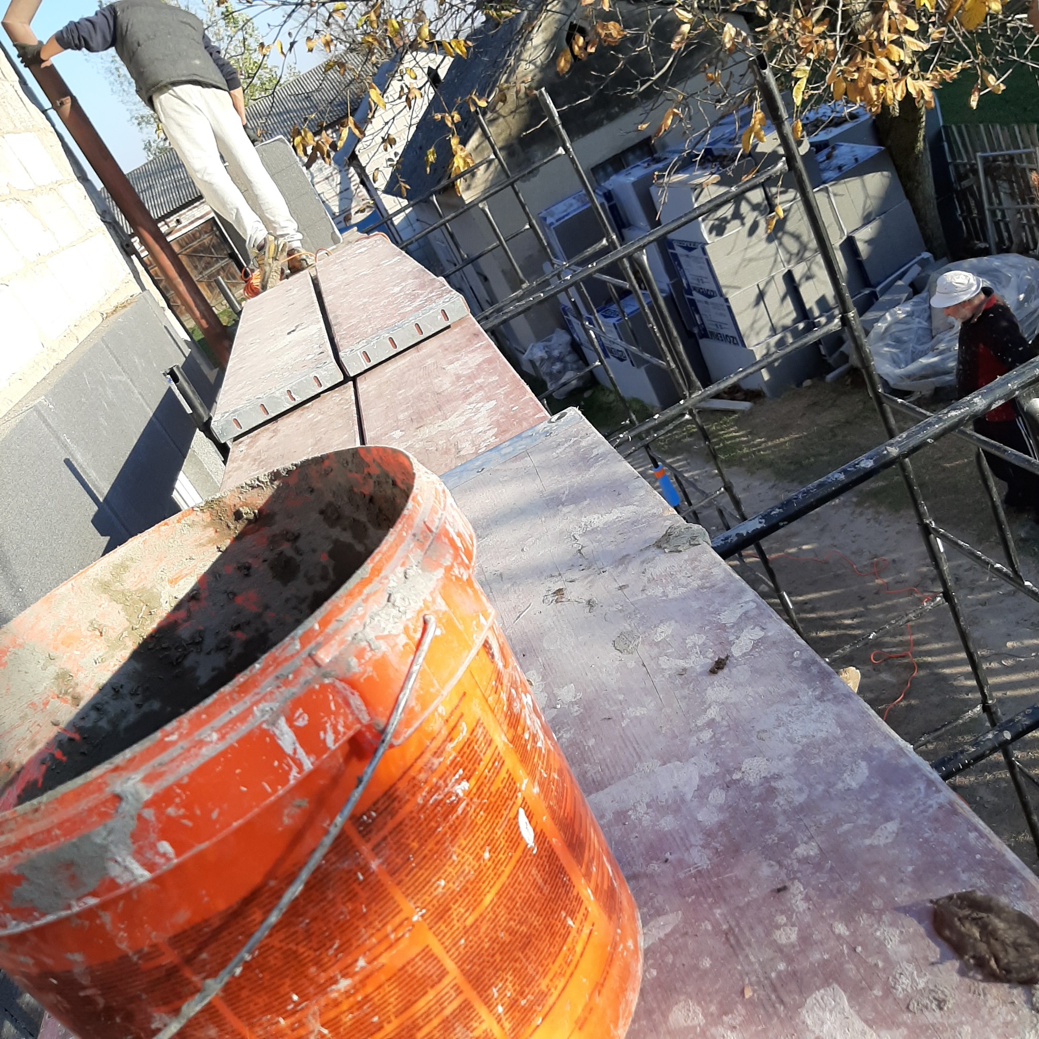 Pomarańczowe wiadro z resztkami zaprawy na drewnianym rusztowaniu, w tle pracownik na rusztowaniu i ocieplone ściany budynku.