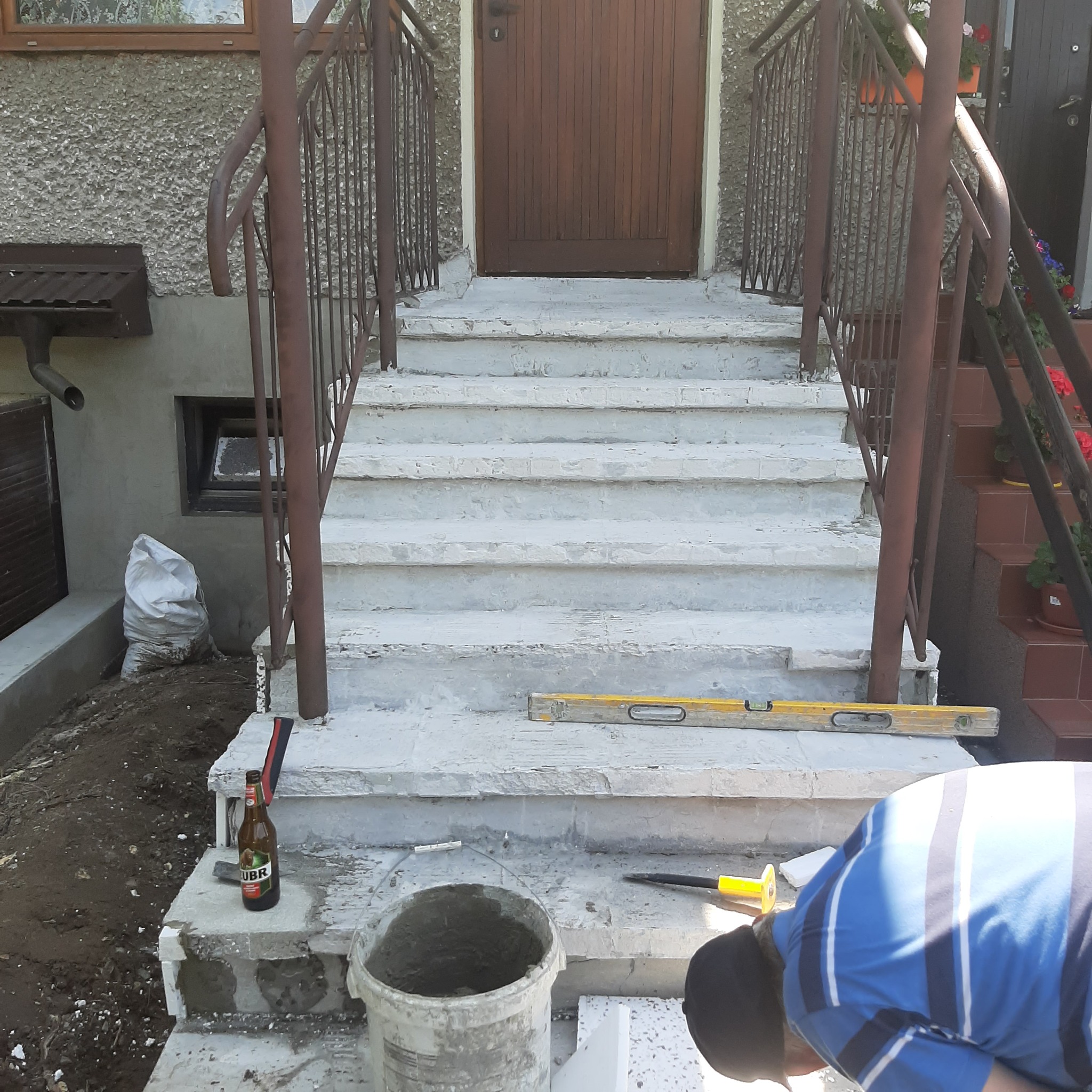 Remont schodów wejściowych: glazurnik układa płytki, widoczna poziomica, wiadro z zaprawą, butelka piwa, schody z brązową barierką, elewacja budynku.
