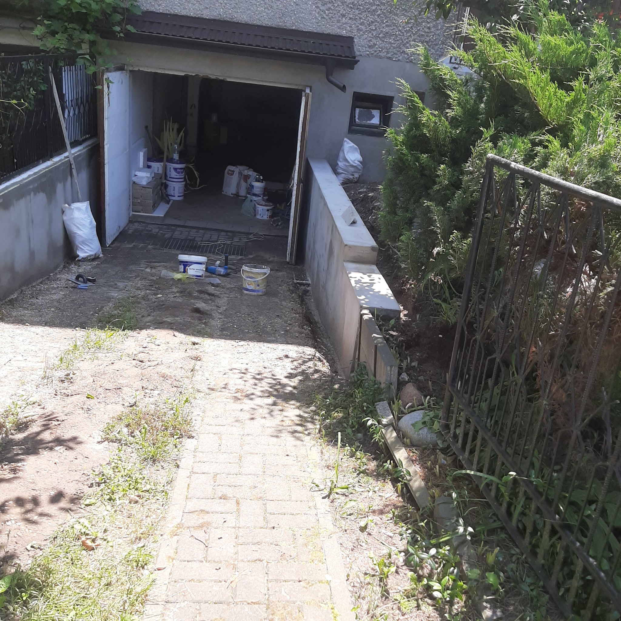 Przygotowania do prac elewacyjnych: otwarty garaż z workami zaprawy, farbami i narzędziami, widok z zewnątrz na ścieżkę prowadzącą do garażu.