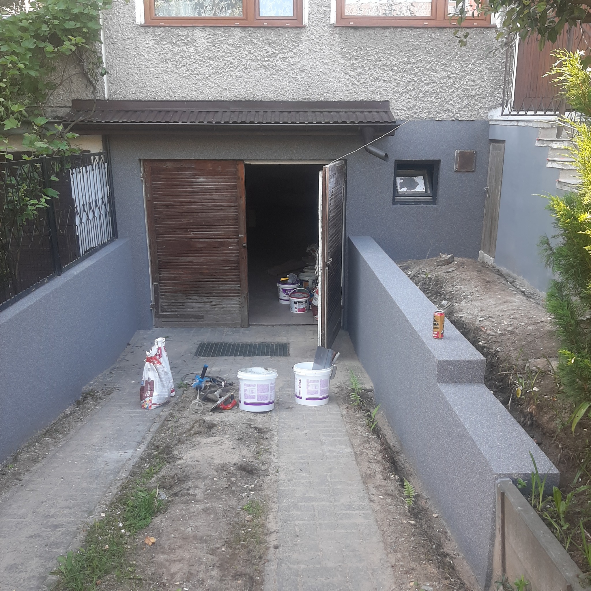 Prace przygotowawcze przed nałożeniem tynku strukturalnego na elewacji budynku z otwartymi drzwiami garażowymi, widoczne wiadra z farbą i narzędzia malarskie.