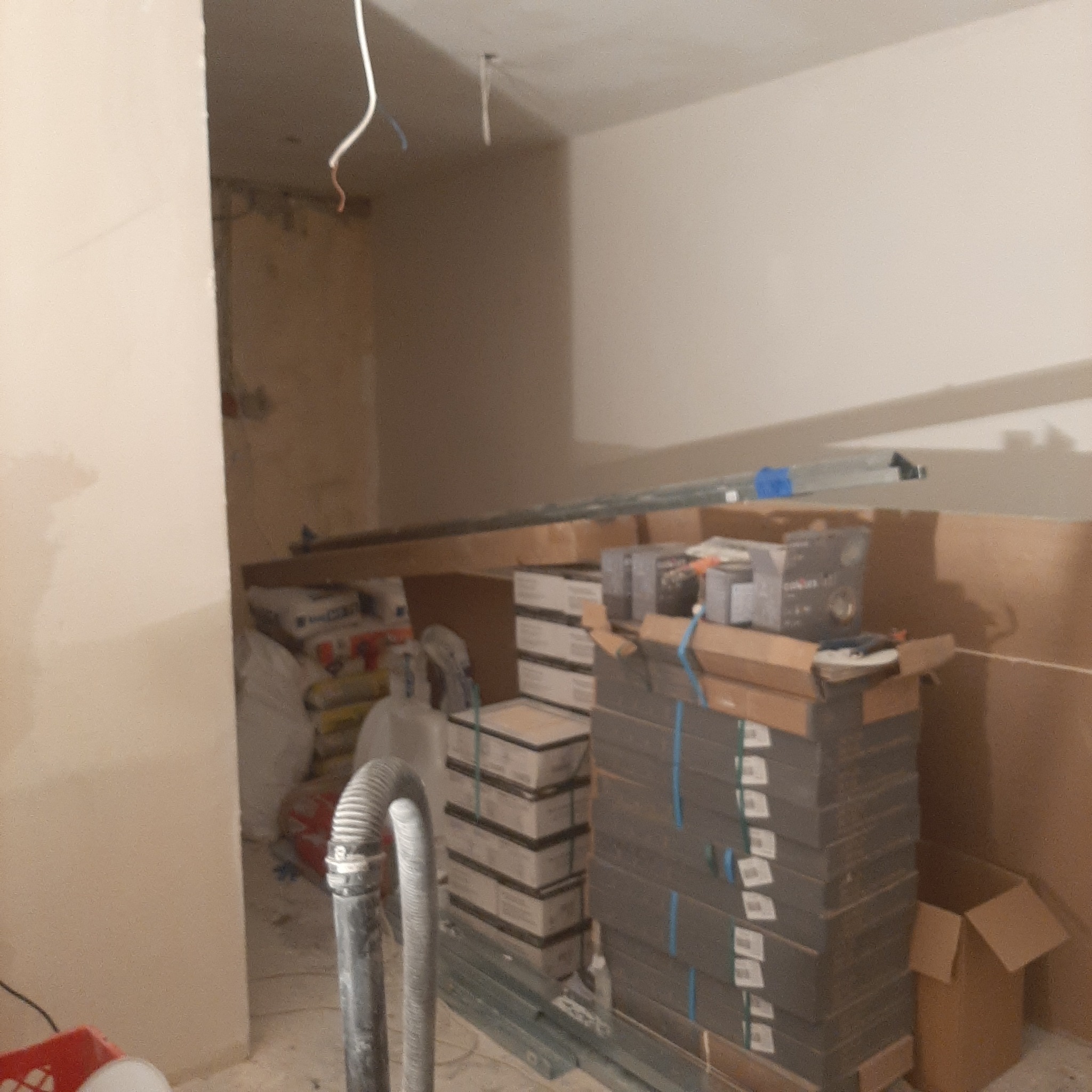 Wnętrze w trakcie remontu: sterta zapakowanych płytek ceramicznych, worki z zaprawą, rura odkurzacza przemysłowego i wystające kable elektryczne.