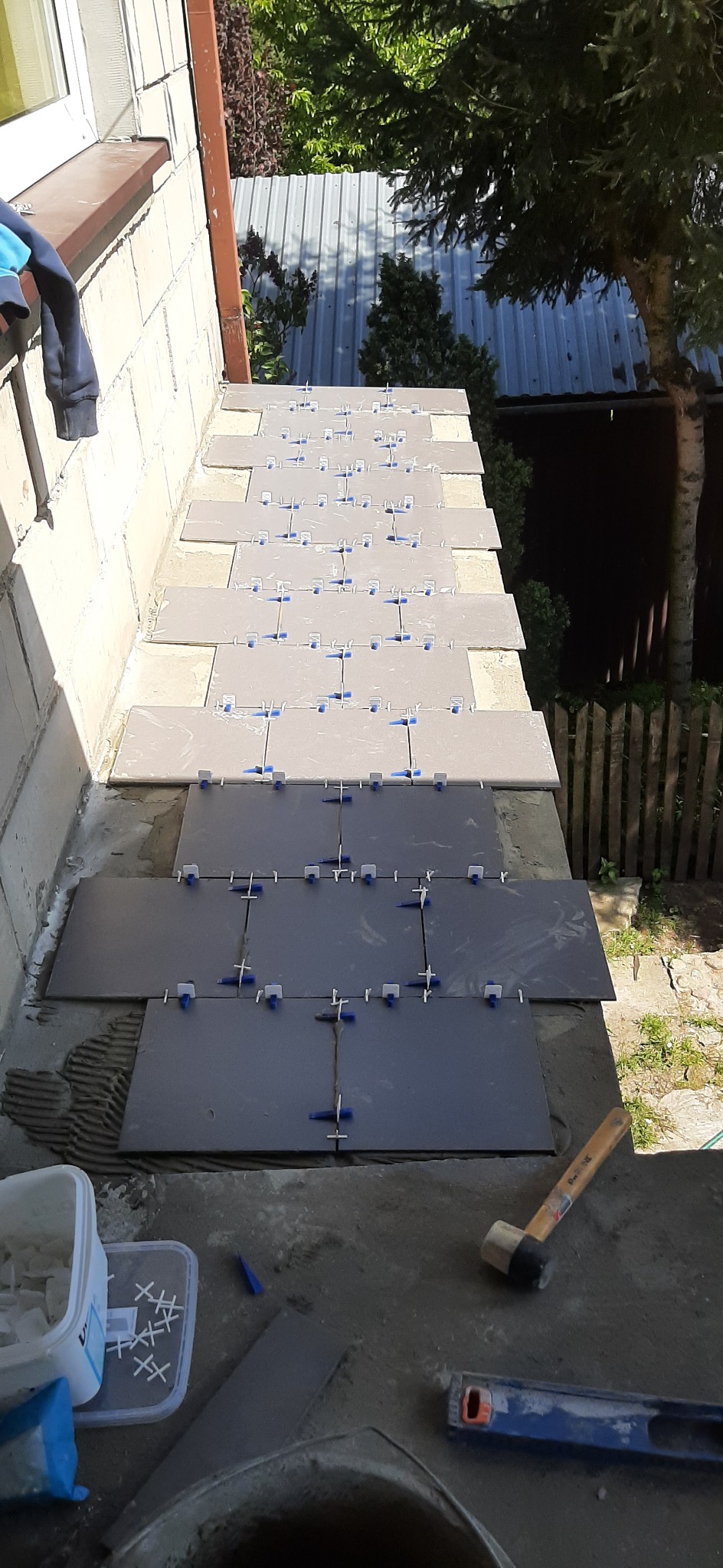 Układanie szarych płytek na balkonie z użyciem krzyżyków dystansowych, widoczny gumowy młotek i poziomica.