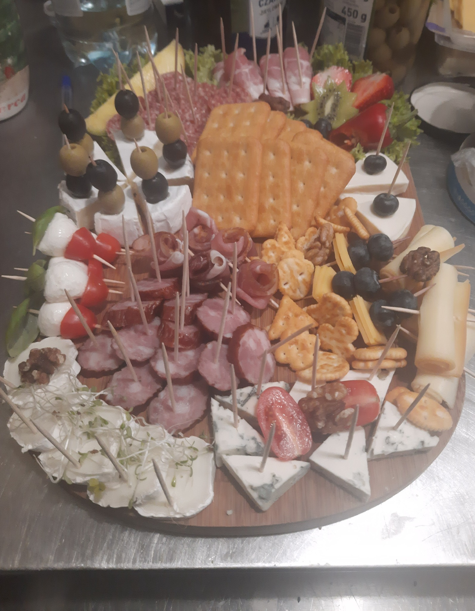 Deska z różnorodnymi przekąskami: sery, wędliny, owoce, oliwki i krakersy, wszystko nabite na wykałaczki, ułożone na drewnianej desce na metalowym blacie.