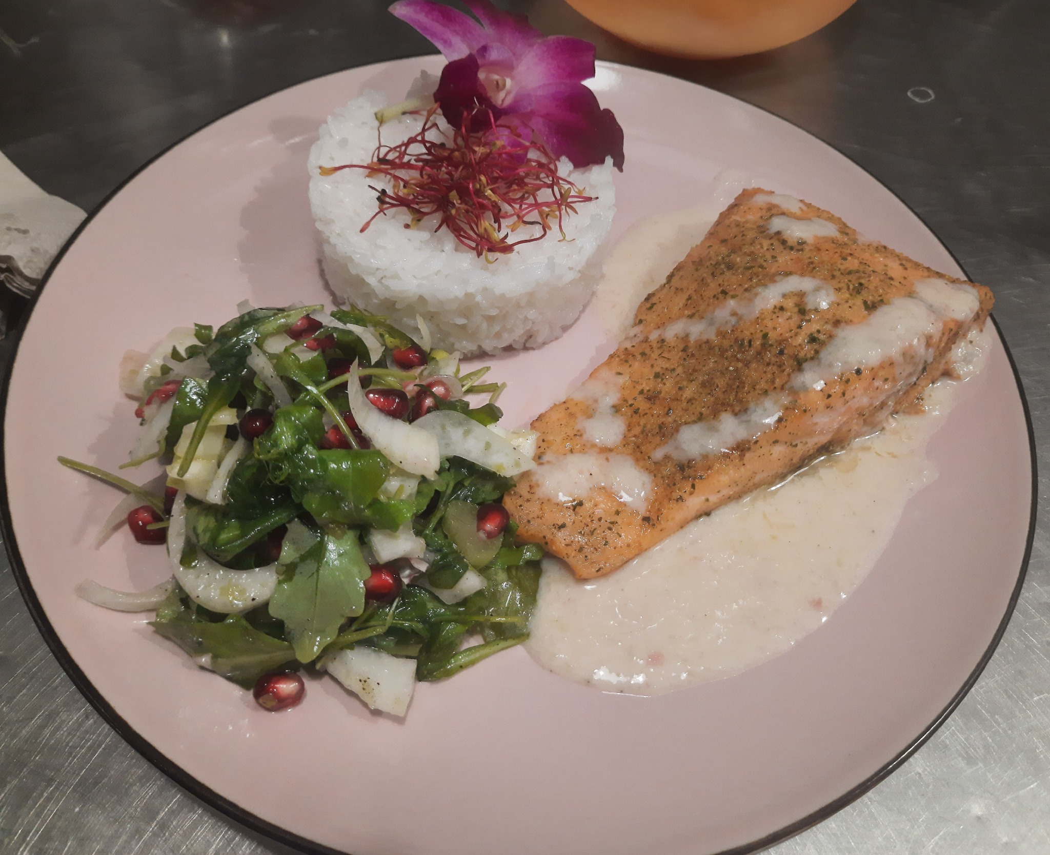 Elegancko podany filet z łososia z sosem, ryżem udekorowanym orchideą i sałatką z granatem na różowym talerzu.