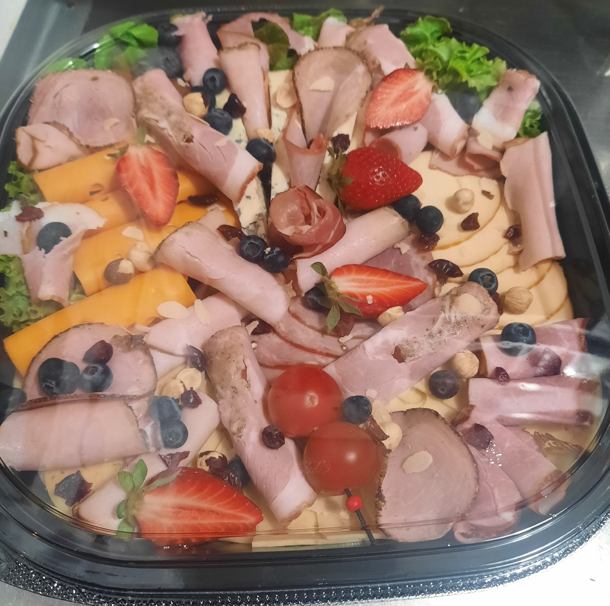 Dekoracyjna taca z wędlinami, serami, truskawkami, borówkami i orzechami laskowymi, ułożona na podkładzie z sałaty.