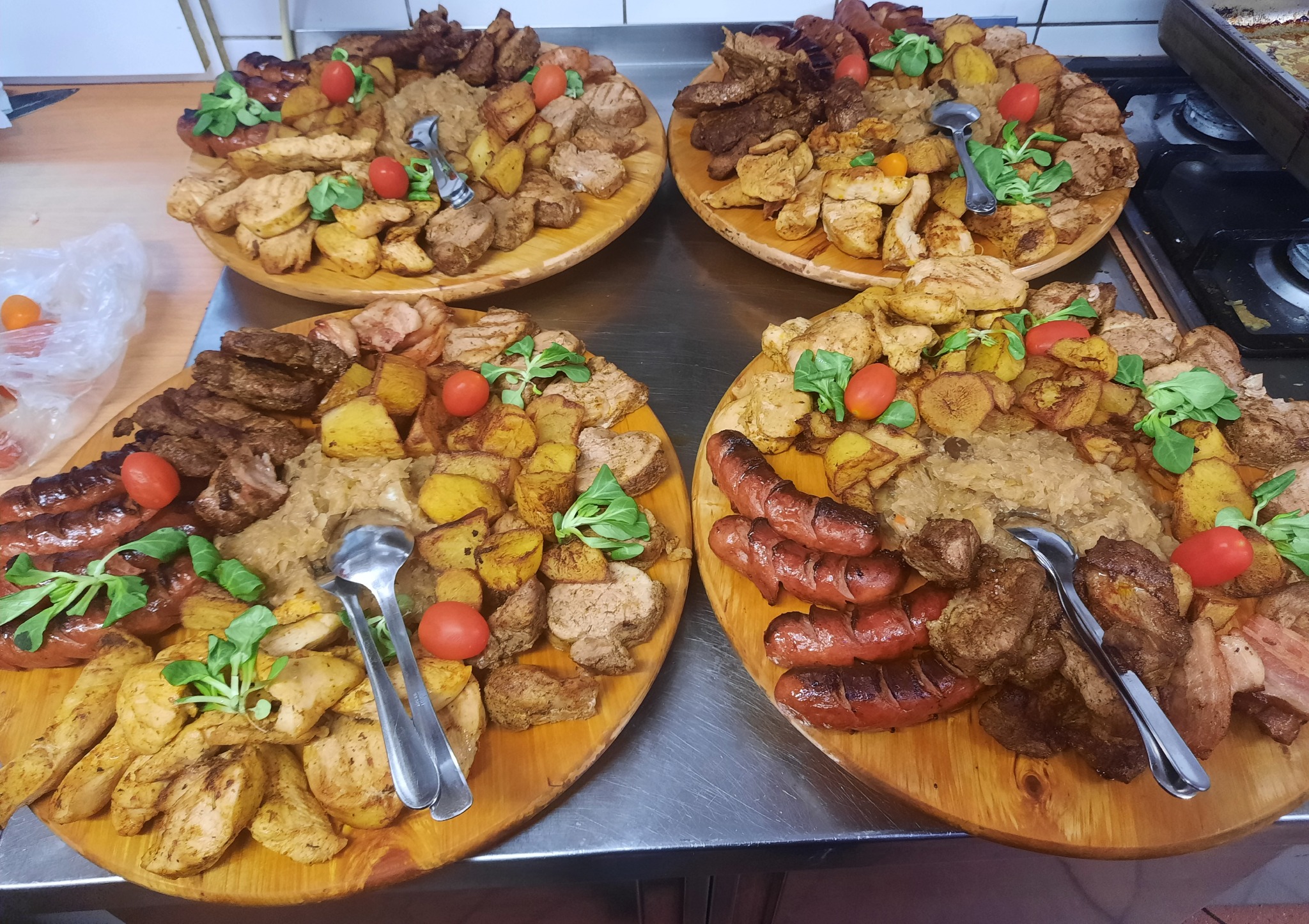 Cztery drewniane tace z różnorodnymi daniami mięsnymi, pieczonymi ziemniakami, kapustą, udekorowane pomidorkami cherry i świeżymi ziołami, srebrne sztućce na każdej tacy.