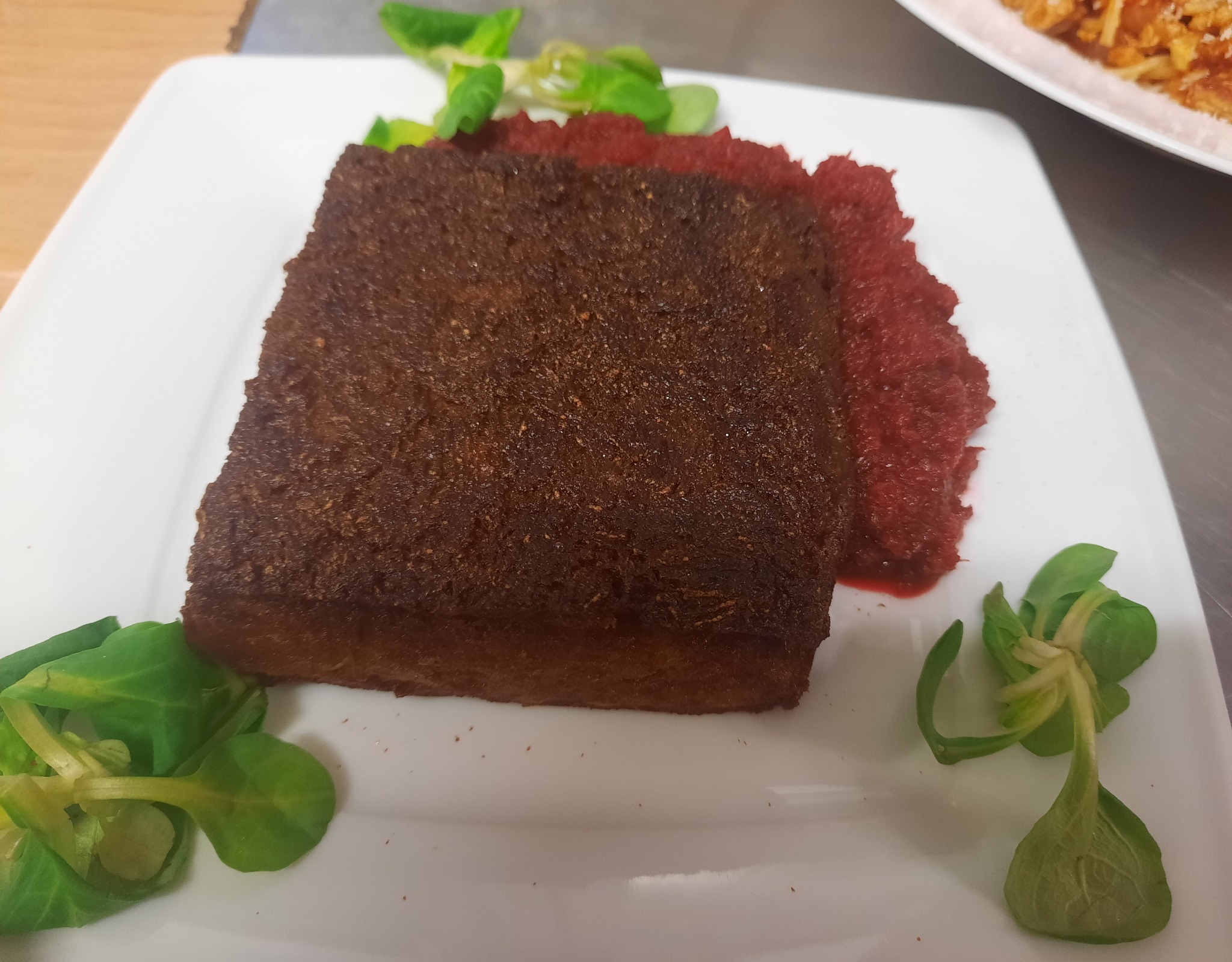 Kwadratowy kawałek pieczonego pasztetu na białym talerzu, udekorowany czerwoną konfiturą i świeżymi listkami sałaty.