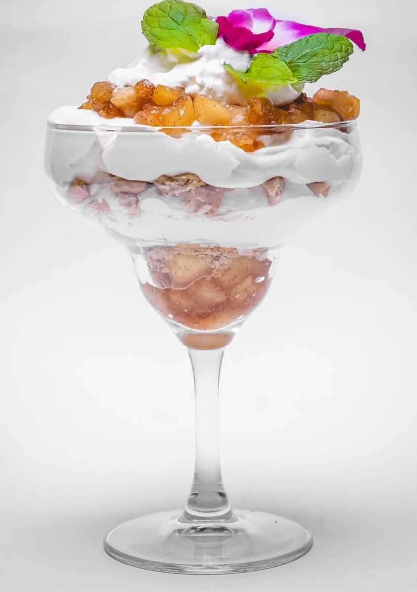 Deser warstwowy w kielichu z jabłkami w karmelu, bitą śmietaną, posypką i dekoracją z mięty oraz różowego kwiatu na białym tle.