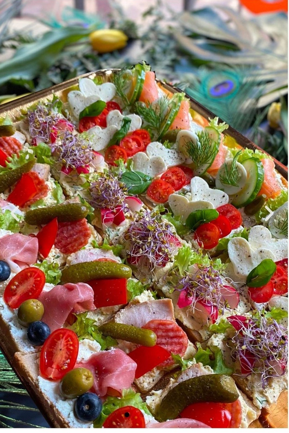 Dekoracyjna taca z kanapkami: serca z mozzarelli, wędzony łosoś, ogórki, pomidorki cherry, salami, szynka, pikle, rzodkiewki i kiełki na jasnym pieczywie, tło z piór pawia.