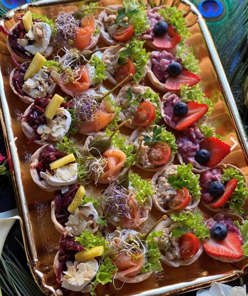 Złota taca z różnorodnymi, apetycznymi tartaletkami: z łososiem i kaparami, z serem i migdałami, z pastą i pomidorem, z owocami jagodowymi i truskawkami, udekorowanymi świeżą sałatą i kiełkami.