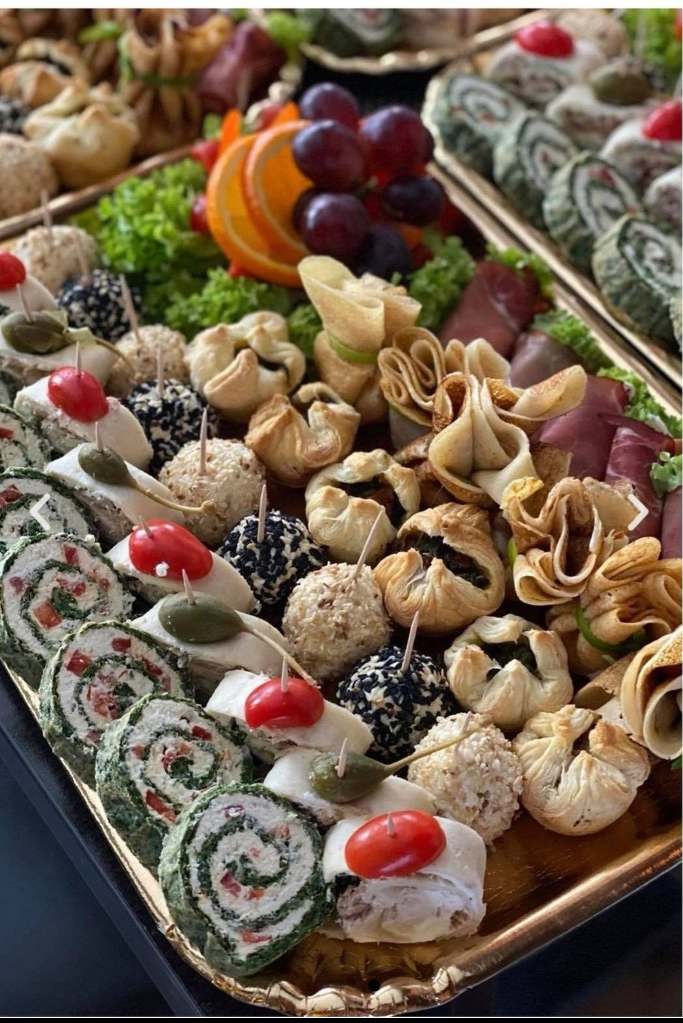 Dekoracyjna złota taca z różnorodnymi przekąskami: roladki szpinakowe, kulki w sezamie i czarnuszce, sakiewki z ciasta francuskiego, plastry wędliny, winogrona i pomarańcze.