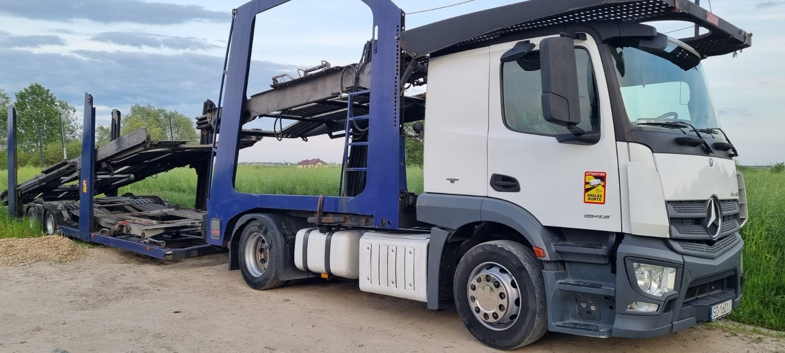 Biała laweta Mercedes z niebieską platformą do przewozu aut, stojąca na szutrowej drodze na tle łąki i błękitnego nieba.