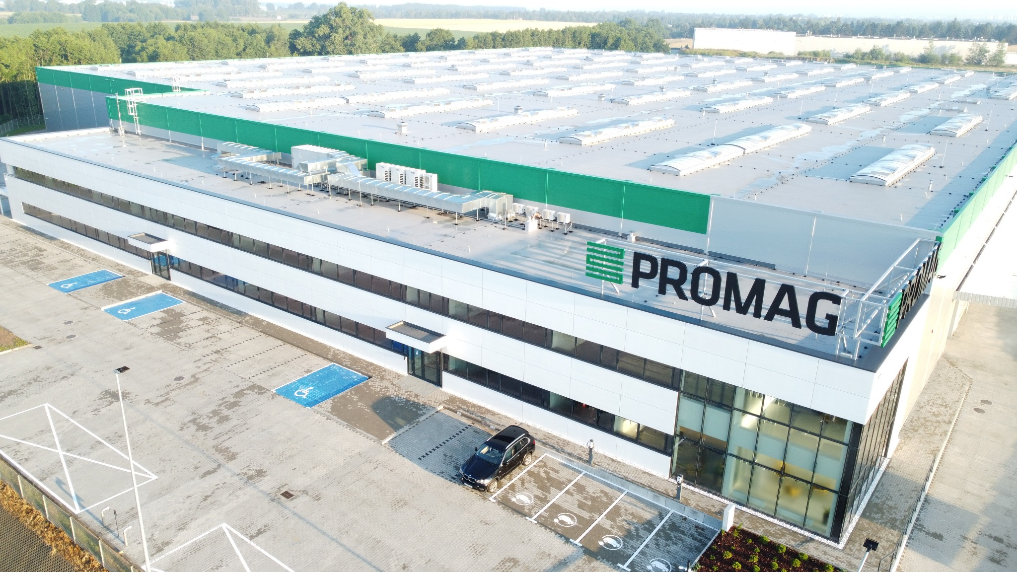 Widok z lotu ptaka na nowoczesny budynek magazynowo-biurowy z zielonym akcentem na dachu i logo firmy PROMAG, z zaparkowanym czarnym SUV-em na parkingu.