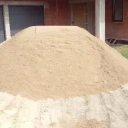 Kupa jasnego materiału sypkiego na ziemi, ślady opon, w tle budynek w trakcie budowy z szarą bramą garażową.