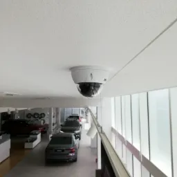 Kopułkowa kamera przemysłowa zamontowana na suficie w salonie samochodowym, widok na rząd pojazdów i przeszkloną ścianę.