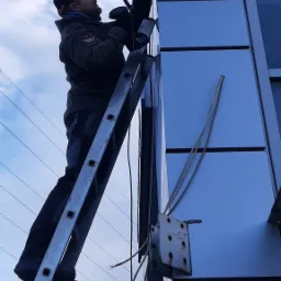 Mężczyzna na drabinie montuje kamerę przemysłową na elewacji budynku, widoczne narzędzia i kable, błękitne niebo w tle.