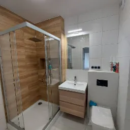 Nowoczesna łazienka z kabiną prysznicową typu walk-in, umywalką nablatową na szafce z drewnopodobnym fornirem i wiszącą miską WC; na półce w kabinie widoczne kosmetyki.