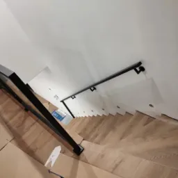 Widok z góry na drewniane schody z czarną metalową balustradą i poręczą przy ścianie, zabezpieczone kartonem na dole.