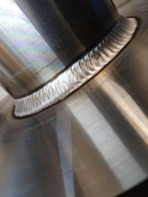 Szczegółowe zbliżenie spawu TIG na nierdzewnej rurze przymocowanej do płaskiej powierzchni, ukazujące regularne, półksiężycowate ślady spoiny.