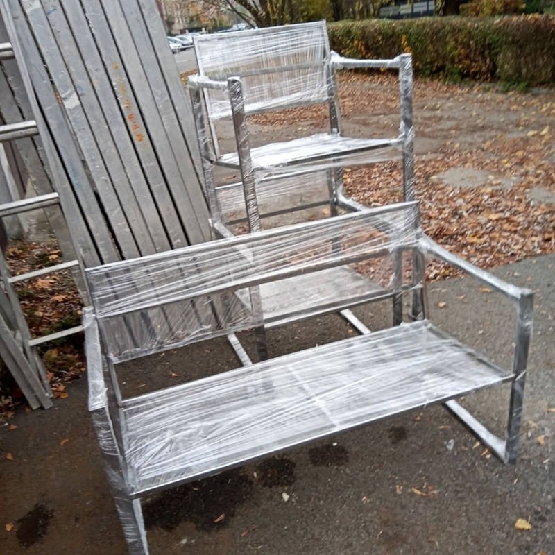 Nowe, metalowe krzesło i ławka ogrodowa, owinięte folią ochronną, ustawione na zewnątrz, obok metalowych profili.