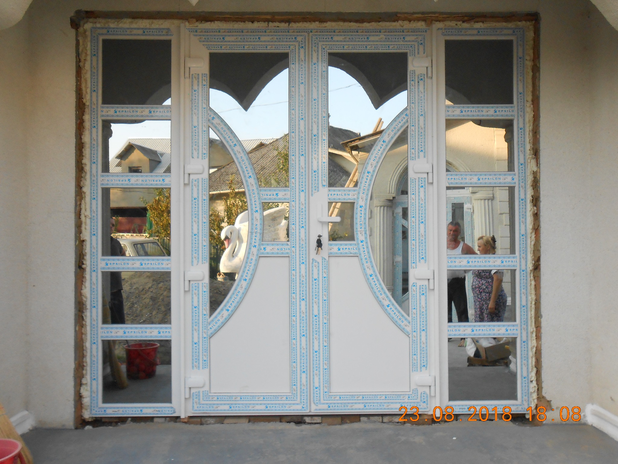 Białe drzwi wejściowe z PCV z przeszkleniami, zamontowane w surowym otworze ściennym, widoczne odbicia otoczenia i osób.