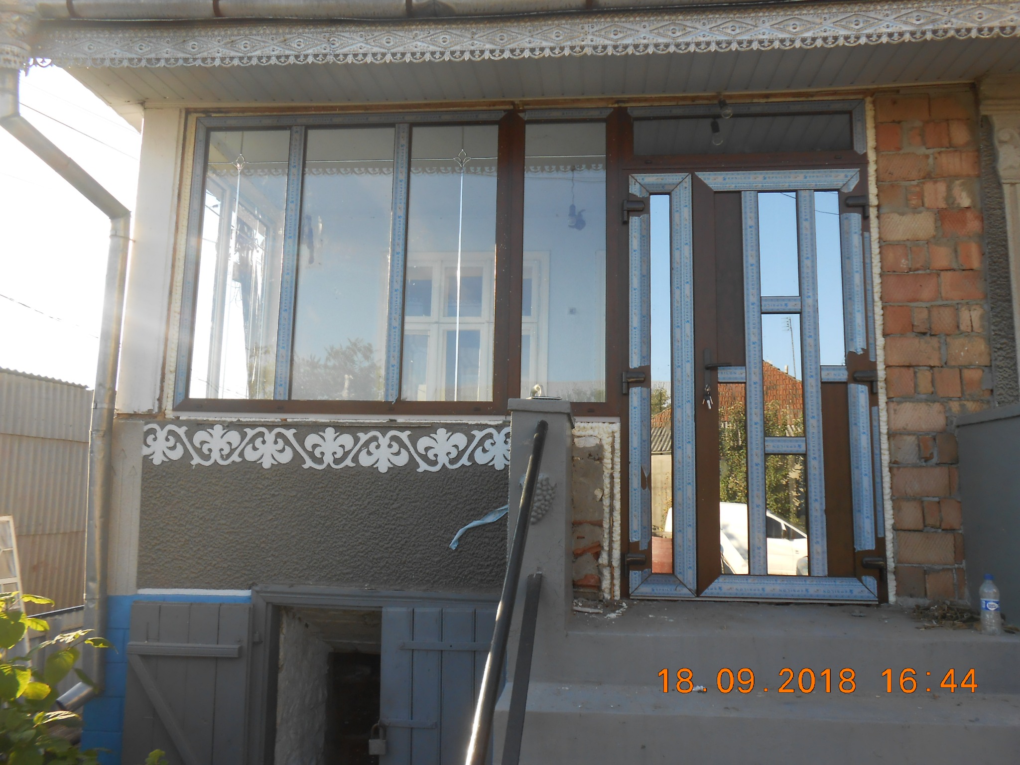 Niedokończony montaż brązowych okien z PCV i drzwi z lustrzanymi wstawkami, widoczne szczeliny wypełnione pianką montażową, na tle ceglanego muru i dekoracyjnego ornamentu na ścianie domu.