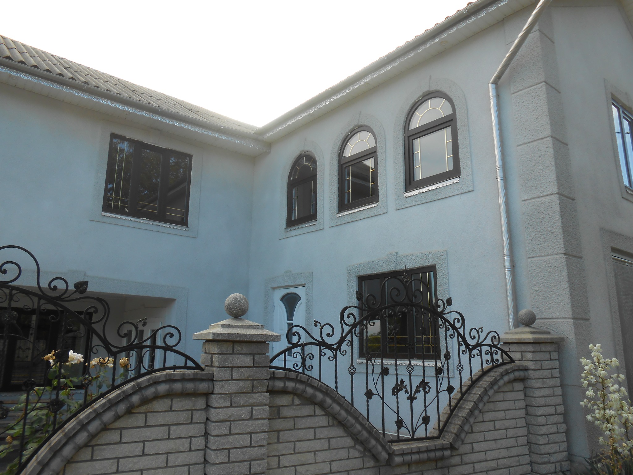 Jasnoszary dom jednorodzinny z ciemnymi oknami, ozdobną bramą z kutego żelaza i ceglanym ogrodzeniem.